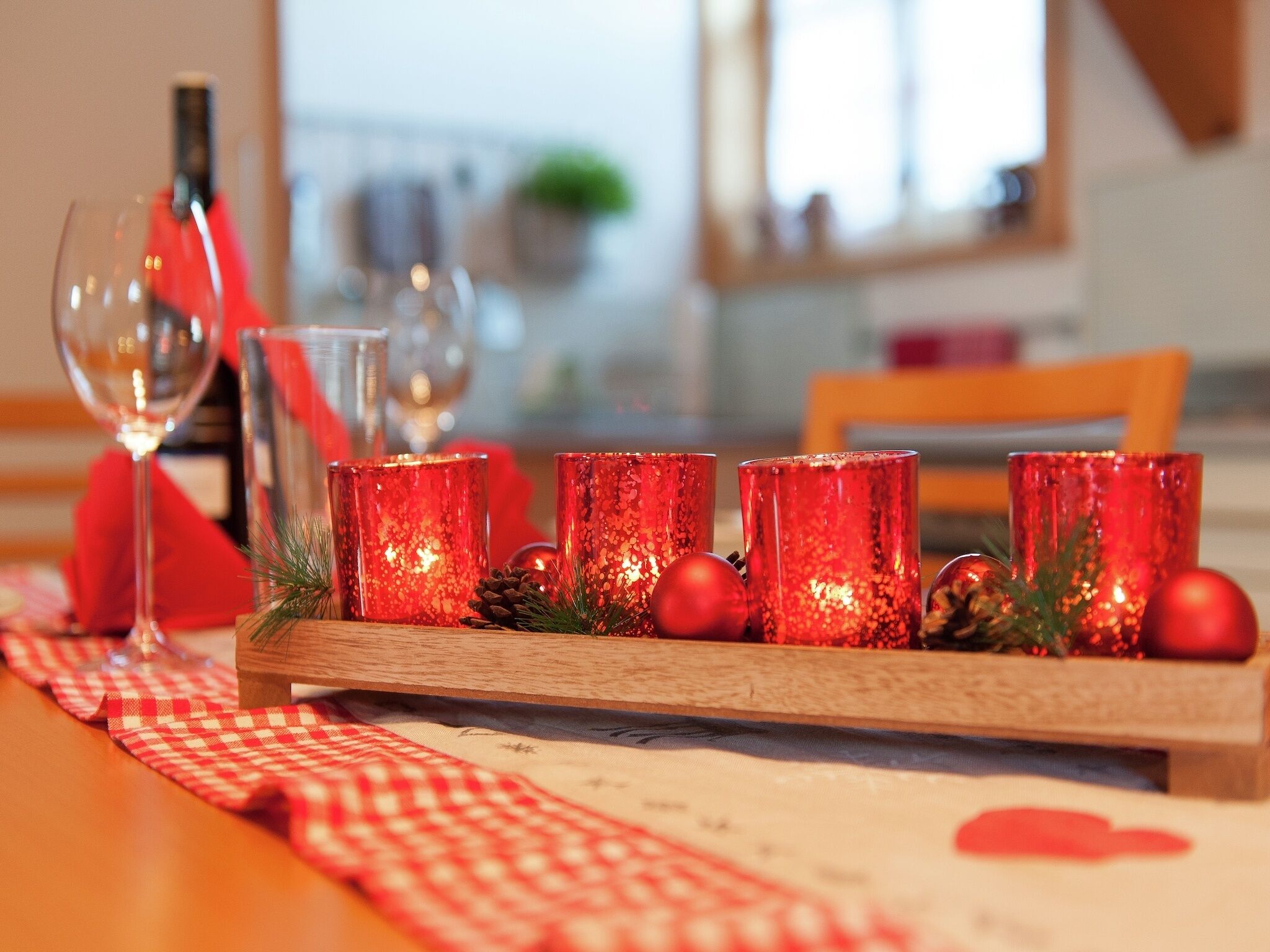 Une table à manger recouverte d’une nappe à carreaux rouges et blancs, avec un verre à vin et un chandelier contenant des bougies rouges et des pommes de pin.
