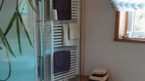 A bathroom with a glass shower enclosure, a towel rack with folded towels, a wooden crate, and a window with blinds.