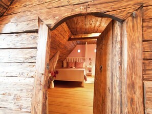 Ein rustikaler Holzraum mit schrägen Wänden, einem Bett mit weißem Kopfteil und einem kleinen Nachttisch mit Blumen in einer Vase.