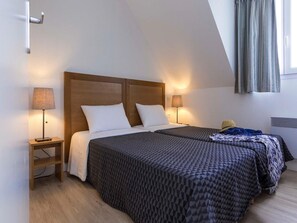 A bedroom with a sloped ceiling, two beds with patterned bedding, bedside lamps, a bedside table, and a window with curtains.