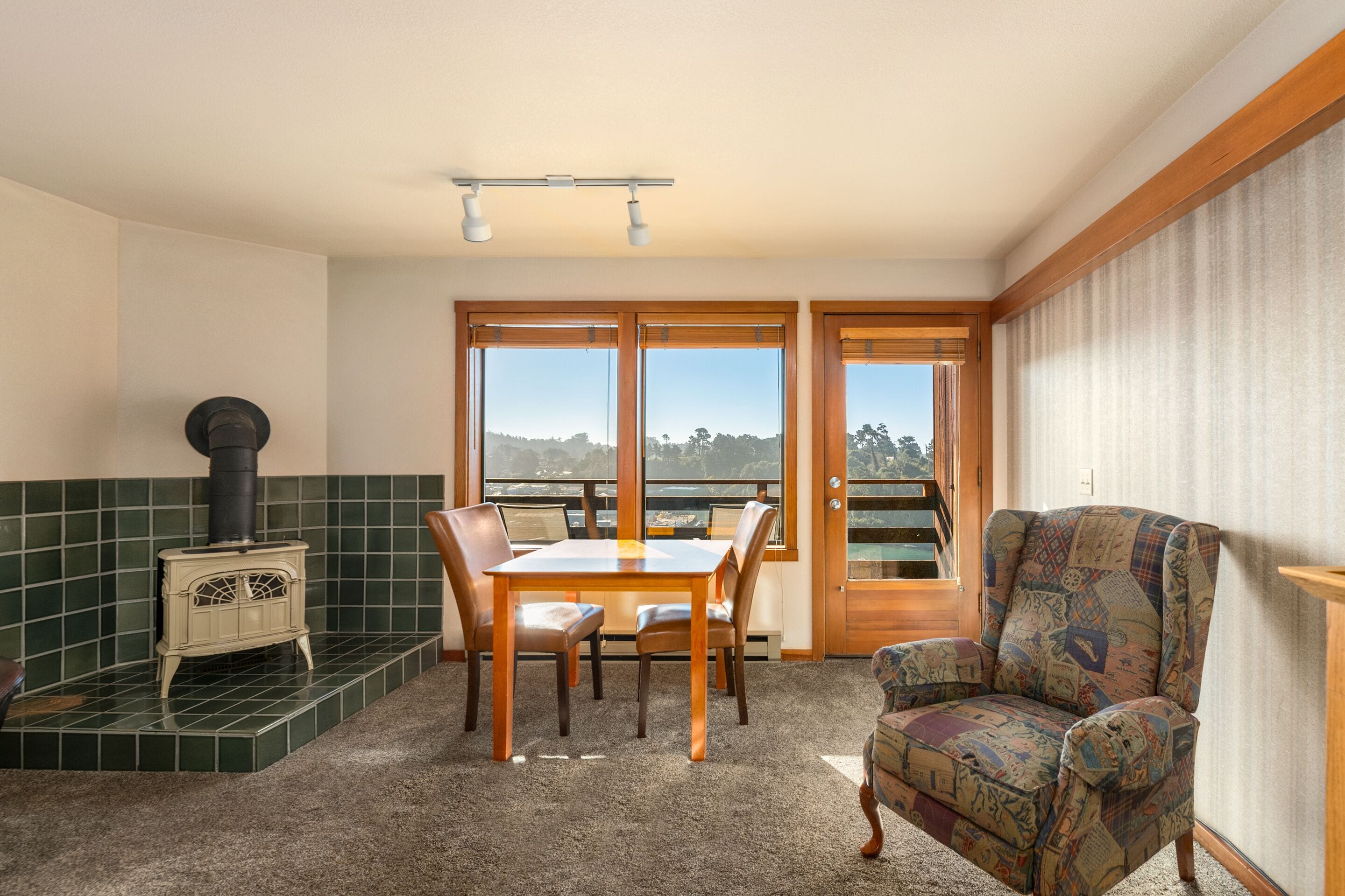 Ein Zimmer mit Esstisch, Stühlen, einem gemusterten Sessel, einem Herd und Blick durch eine Glastür.