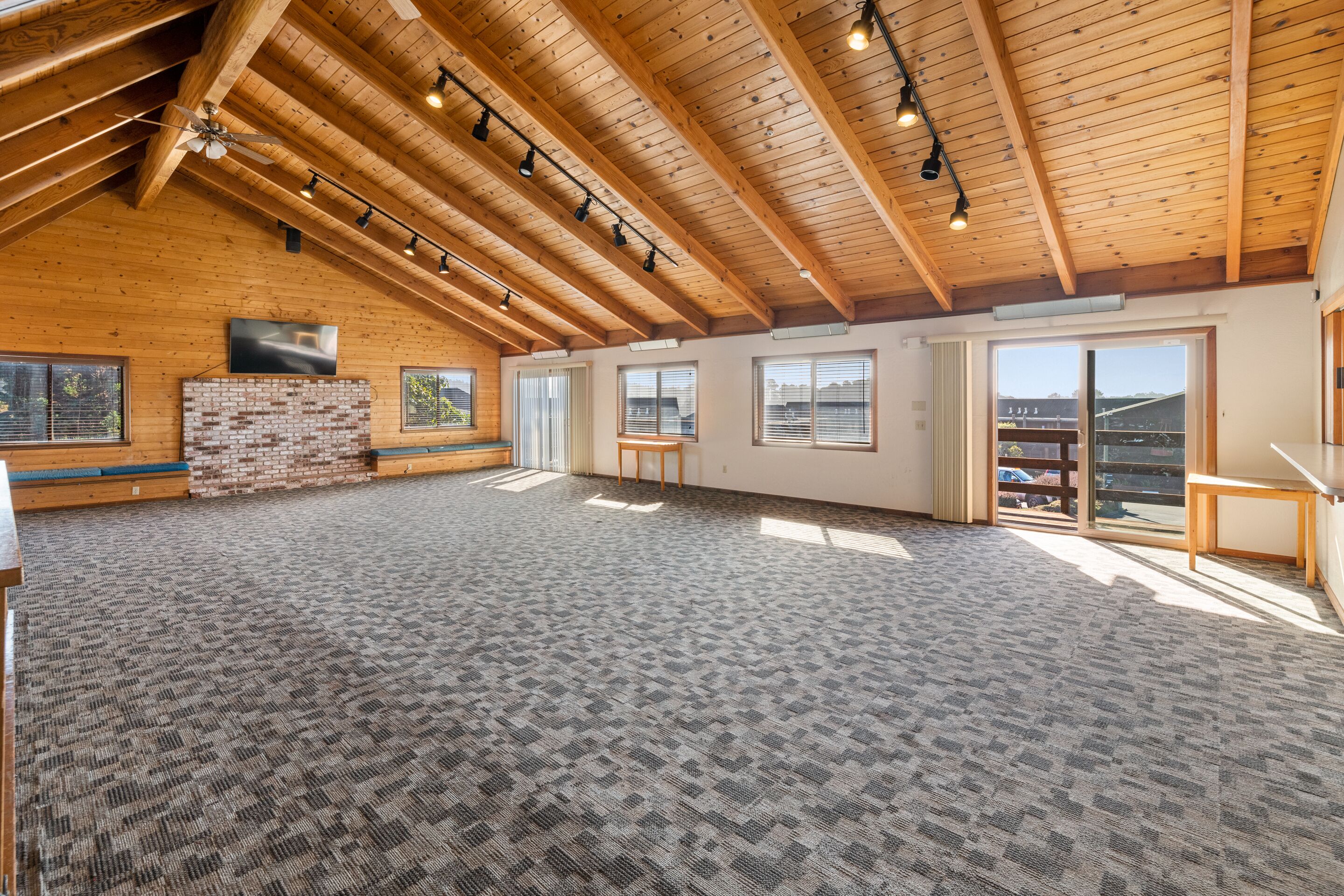 A spacious room with a high ceiling, wooden beams, a brick fireplace, large windows, and a carpeted floor.