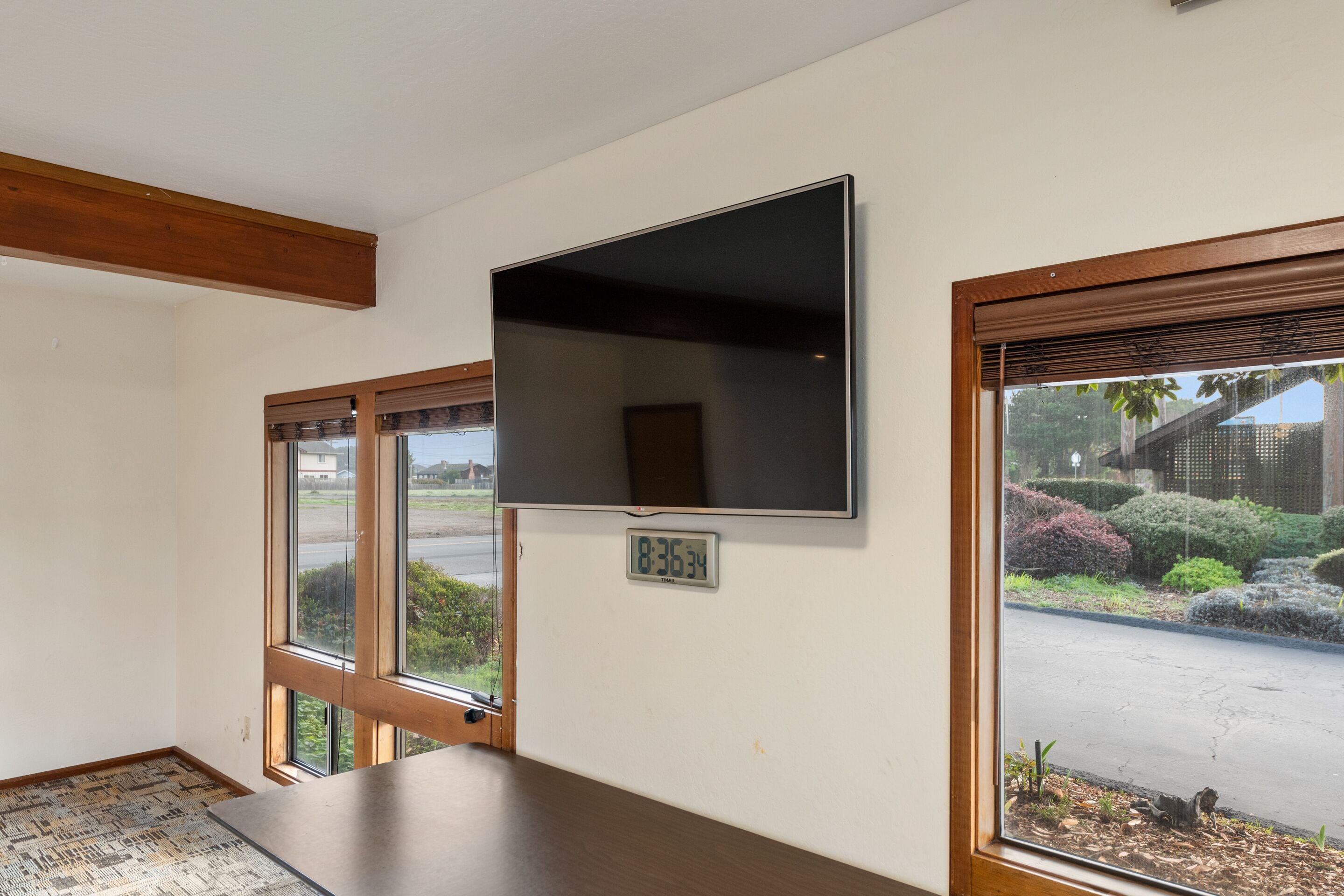 A room with a large flat-screen TV mounted on the wall, a wooden table, and a window with a view of a street and greenery.