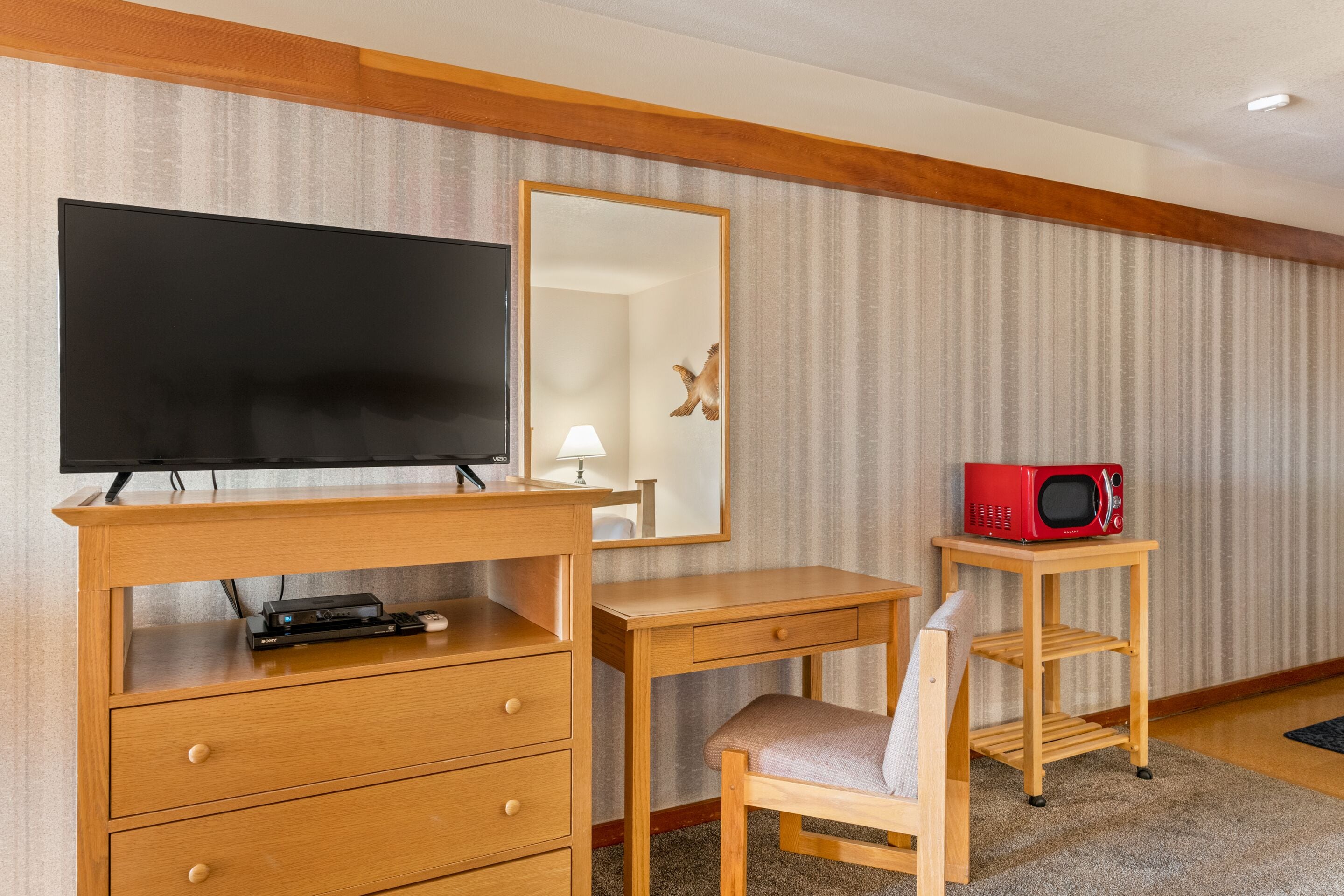 A hotel room with a wooden desk, a television, a red microwave, a mirror, and a chair.