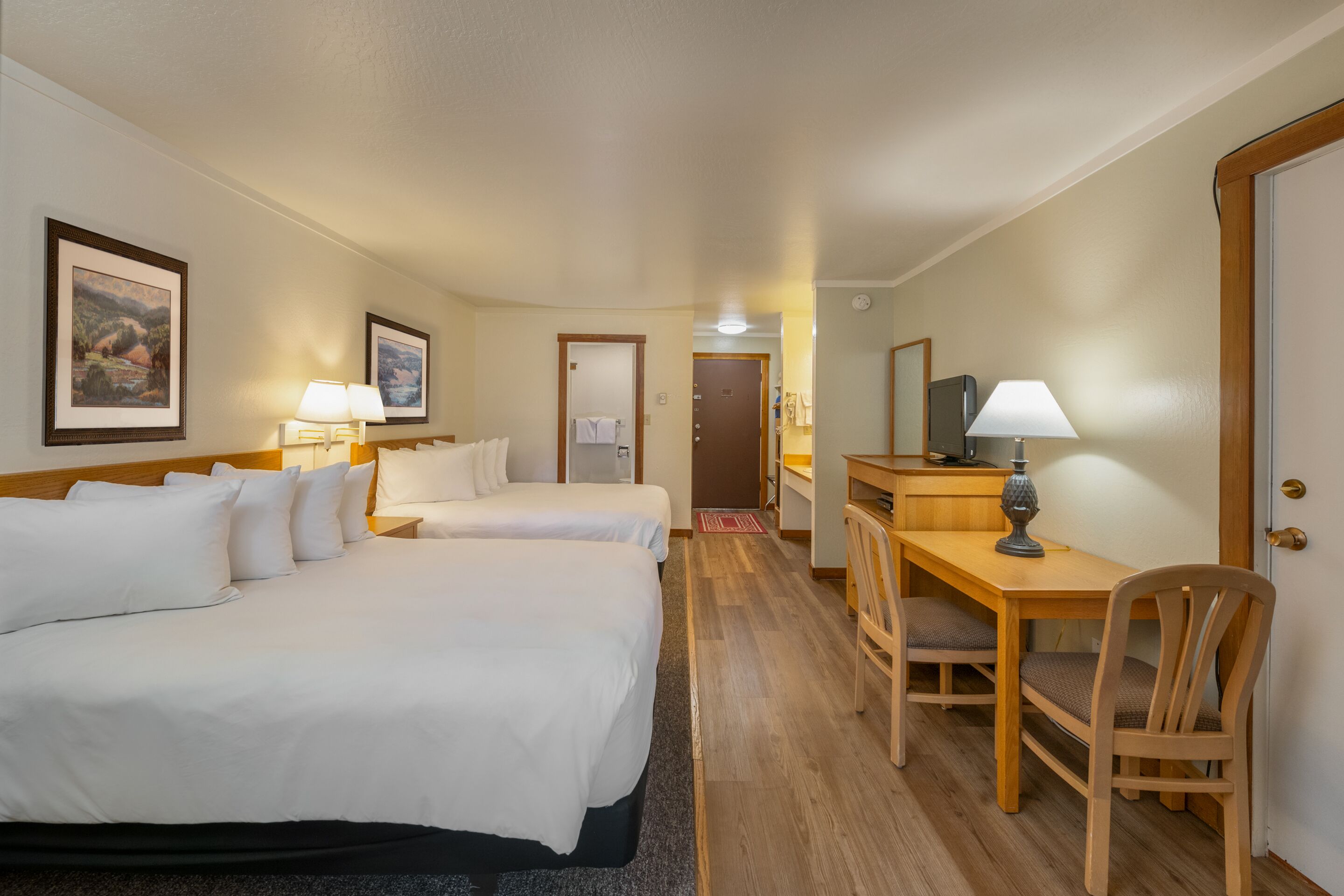 A hotel room with two beds, a desk, a chair, and a lamp.