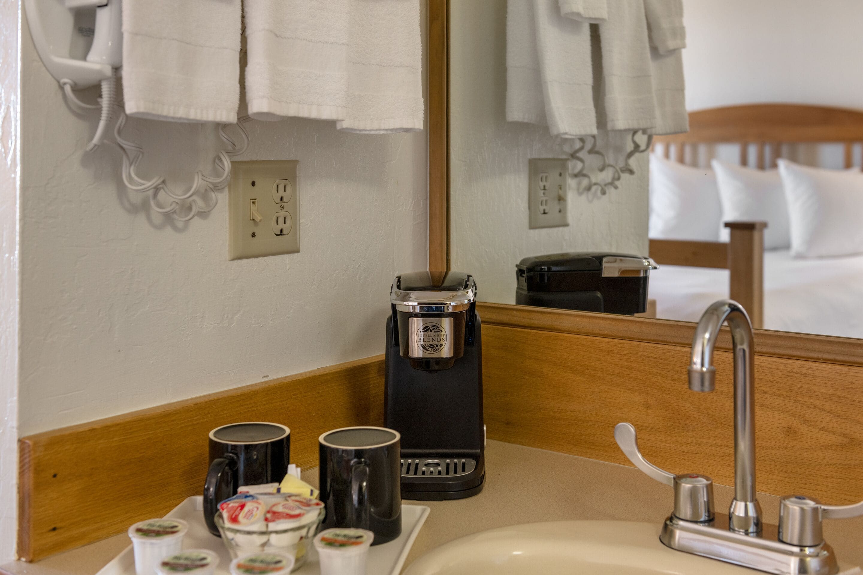 Un baño de hotel con lavamanos, cafetera y espejo que refleja una cama y toallas.