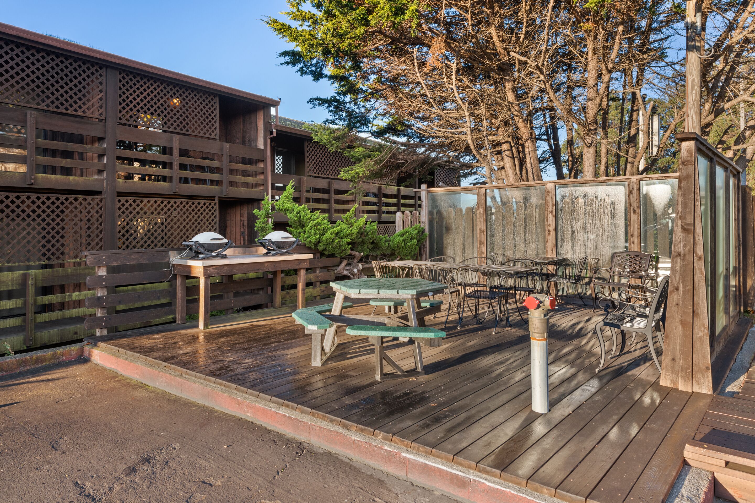 A wooden deck with picnic tables and chairs, a grill, and a fenced area with trees.