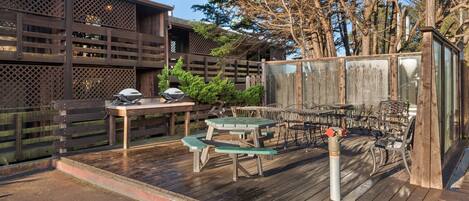 A wooden deck with picnic tables and chairs, a grill, and a fenced area with trees.