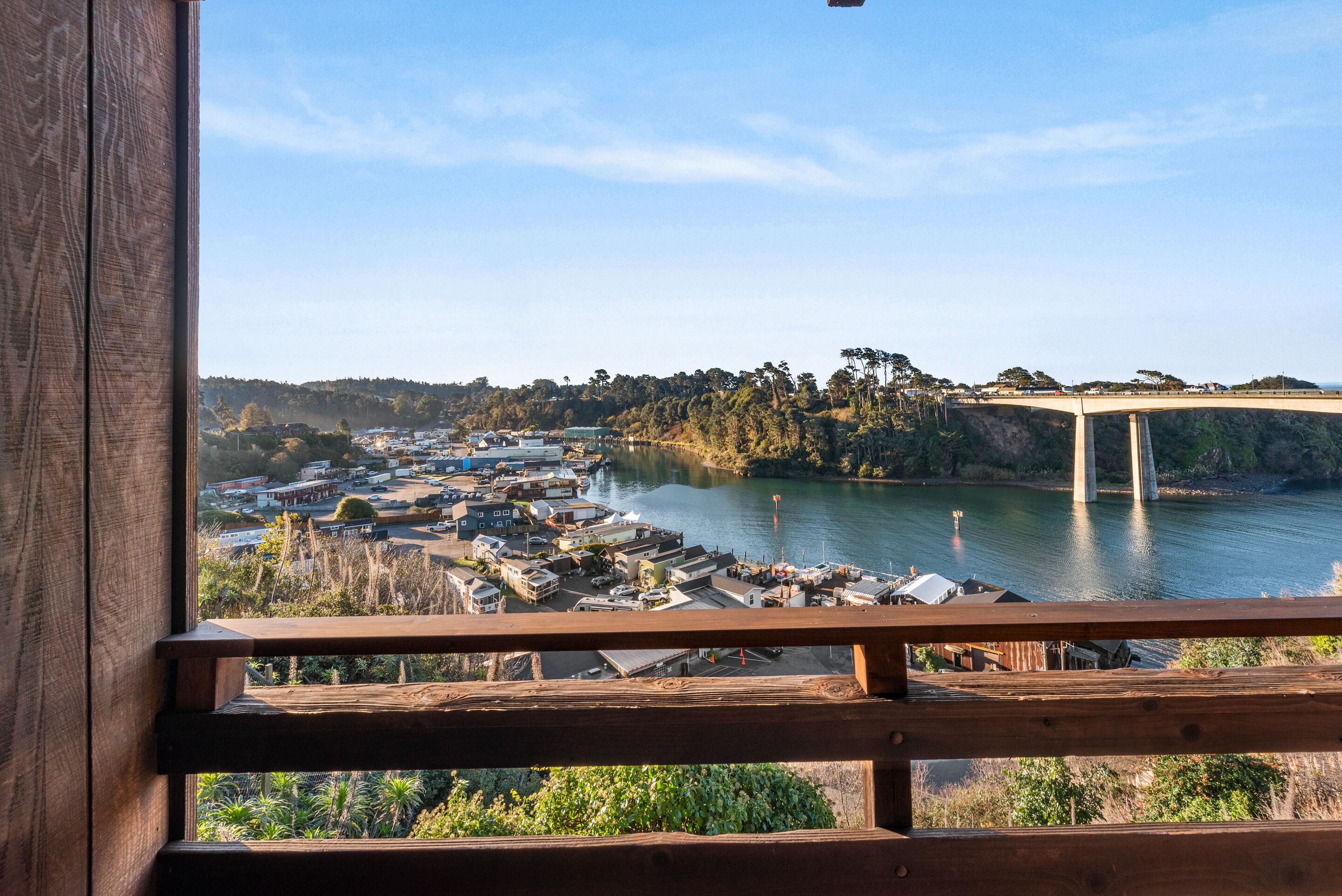 A view from a wooden balcony overlooking a riverside town with a bridge, buildings, and greenery.
