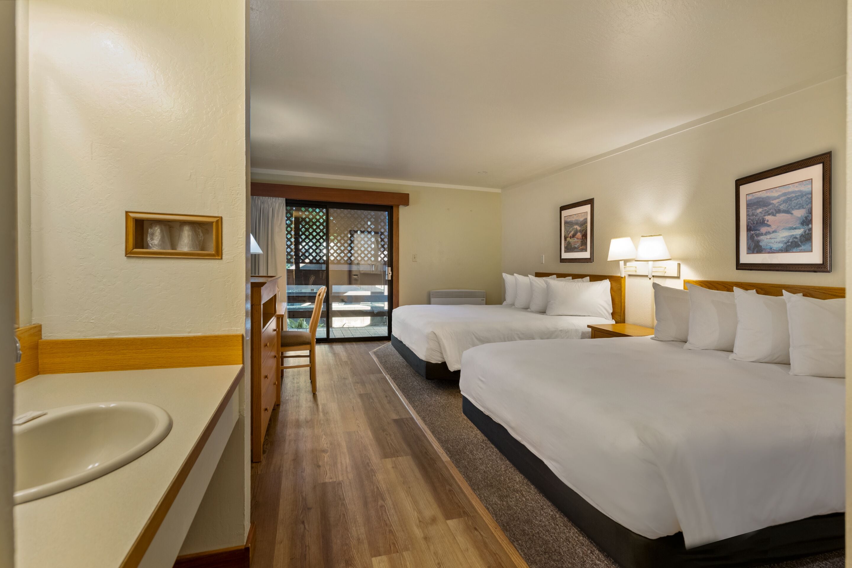 A hotel room with two beds, a bathroom with a sink, and a view through a sliding door to a balcony.