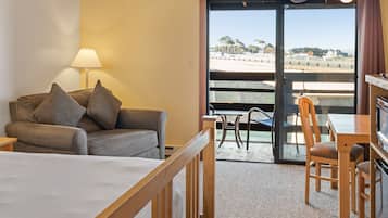 A hotel room with a bed, a sofa, a table, and a chair. There is a view of the beach through the glass door.