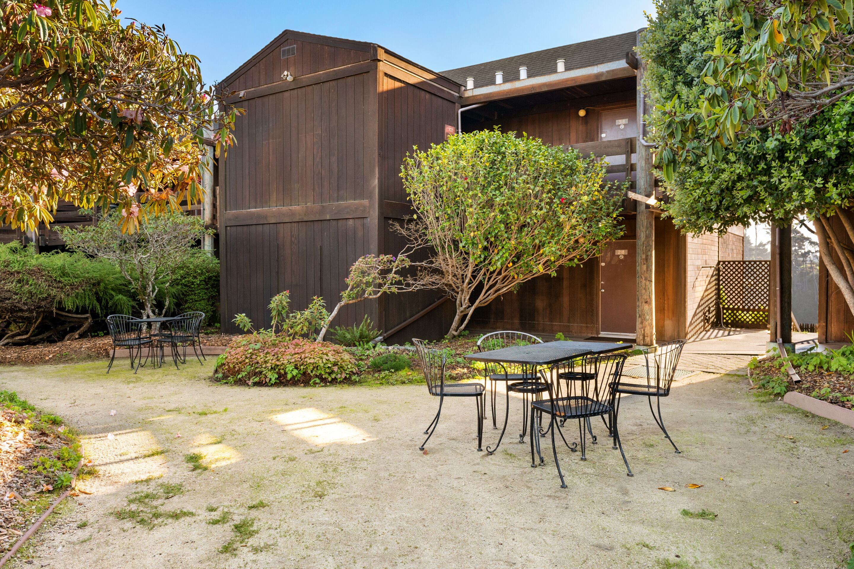 A patio with a table and chairs, surrounded by greenery and a wooden building.