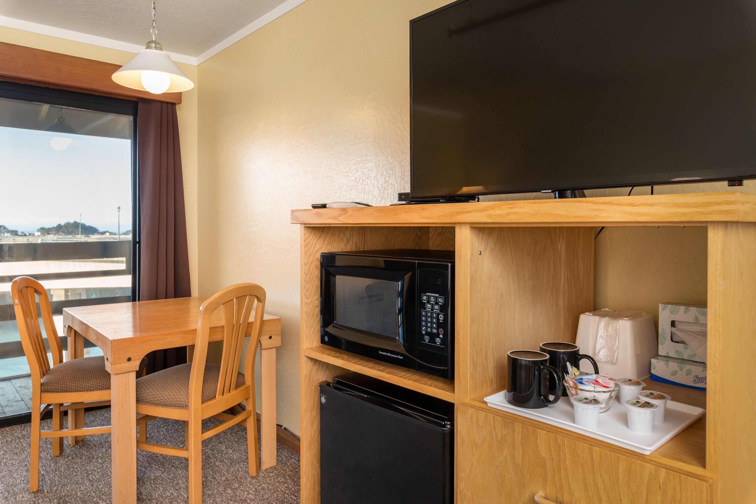 A compact hotel room with a wooden dining table, two chairs, a microwave, a small refrigerator, a flat-screen TV, and a tray with cups and a box of tissues.