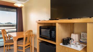 A compact hotel room with a wooden dining table, two chairs, a microwave, a small refrigerator, a flat-screen TV, and a tray with cups and a box of tissues.