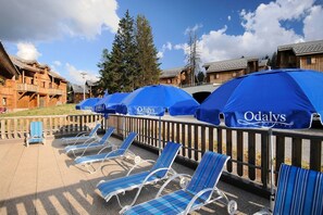Gemütliche Liegestühle mit blau gestreiften Kissen und Sonnenschirme mit 'Odalys'-Branding, vor einer Kulisse aus Holzbauten und blauem Himmel.