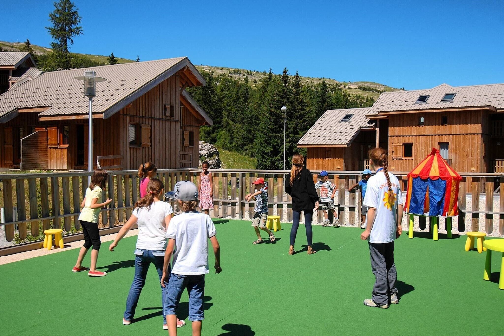 A group of people on a green outdoor area with wooden cabins in the background.