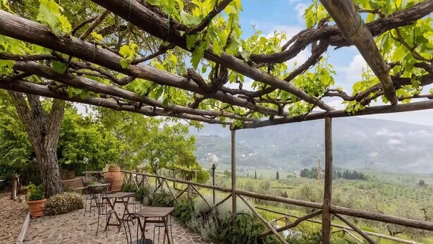 Un espacio de estar al aire libre rústico con suelo de piedra, mesas y sillas de madera, con vistas a un valle pintoresco.