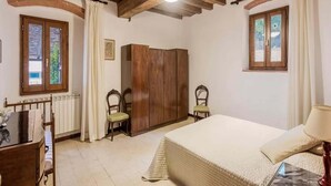 Un dormitorio con techo de madera, una cama con edredón blanco, dos sillas, un armario de madera y una ventana con cortinas.