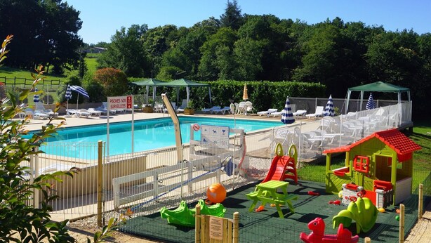Une piscine entourée d’une clôture, une aire de jeux pour enfants et un petit bâtiment avec un toit rouge.