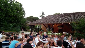 Un espace extérieur aménagé pour les repas, avec des personnes assises à des tables, entourées de verdure et d’un bâtiment doté d’un toit en tuiles.