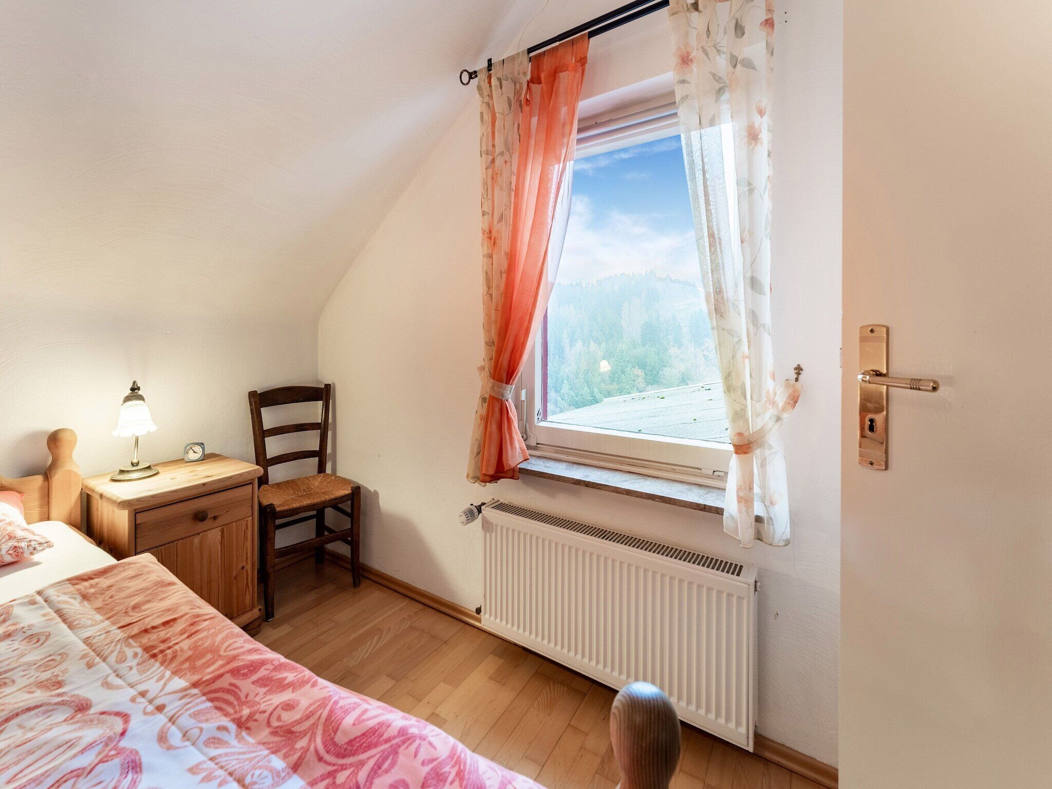 A bedroom with a bed, a nightstand with a lamp, a chair, a window with curtains, a radiator, and a door with a handle.