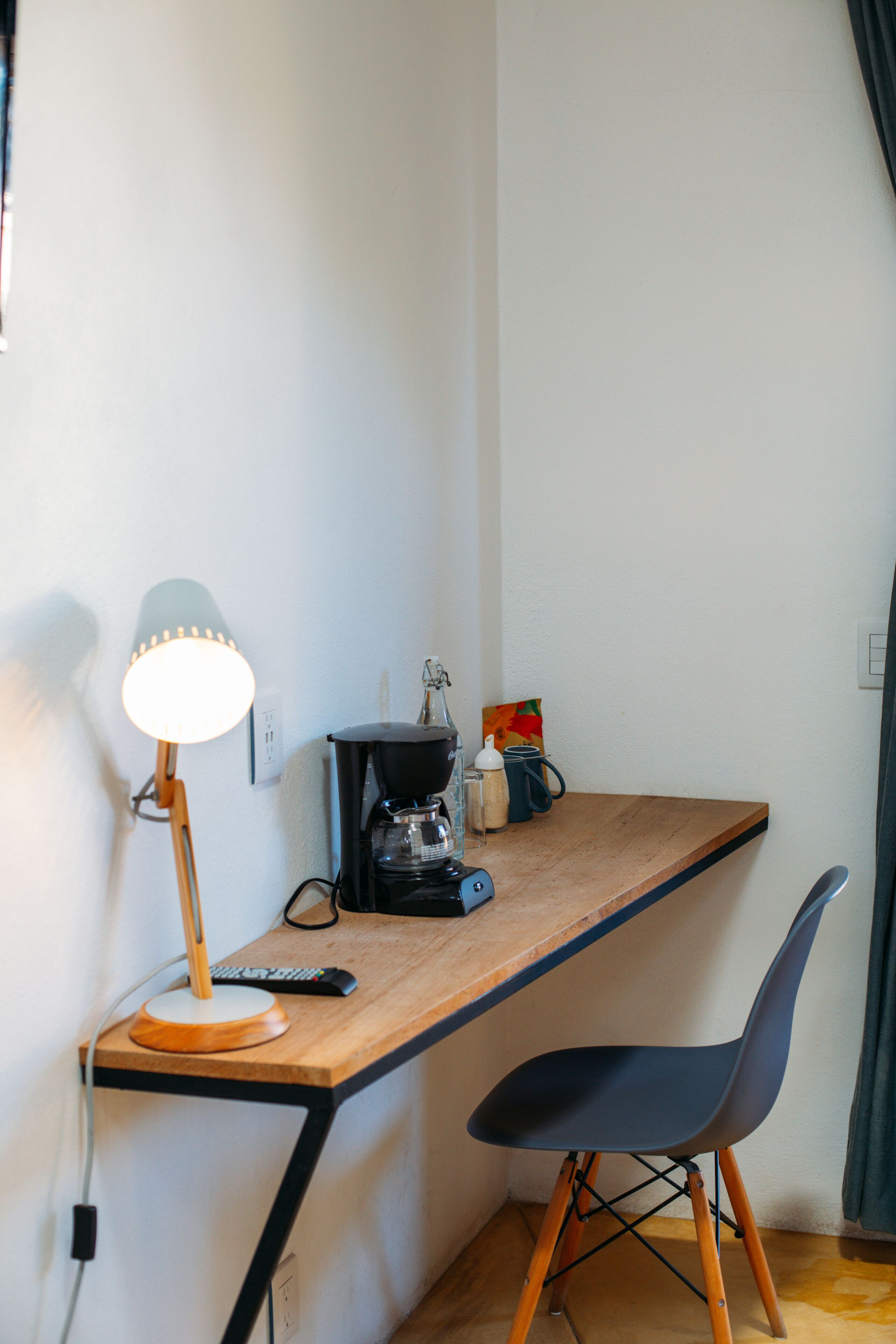 Un petit espace de travail avec un bureau en bois, une chaise noire, une lampe, une machine à café et un tableau accroché au mur.