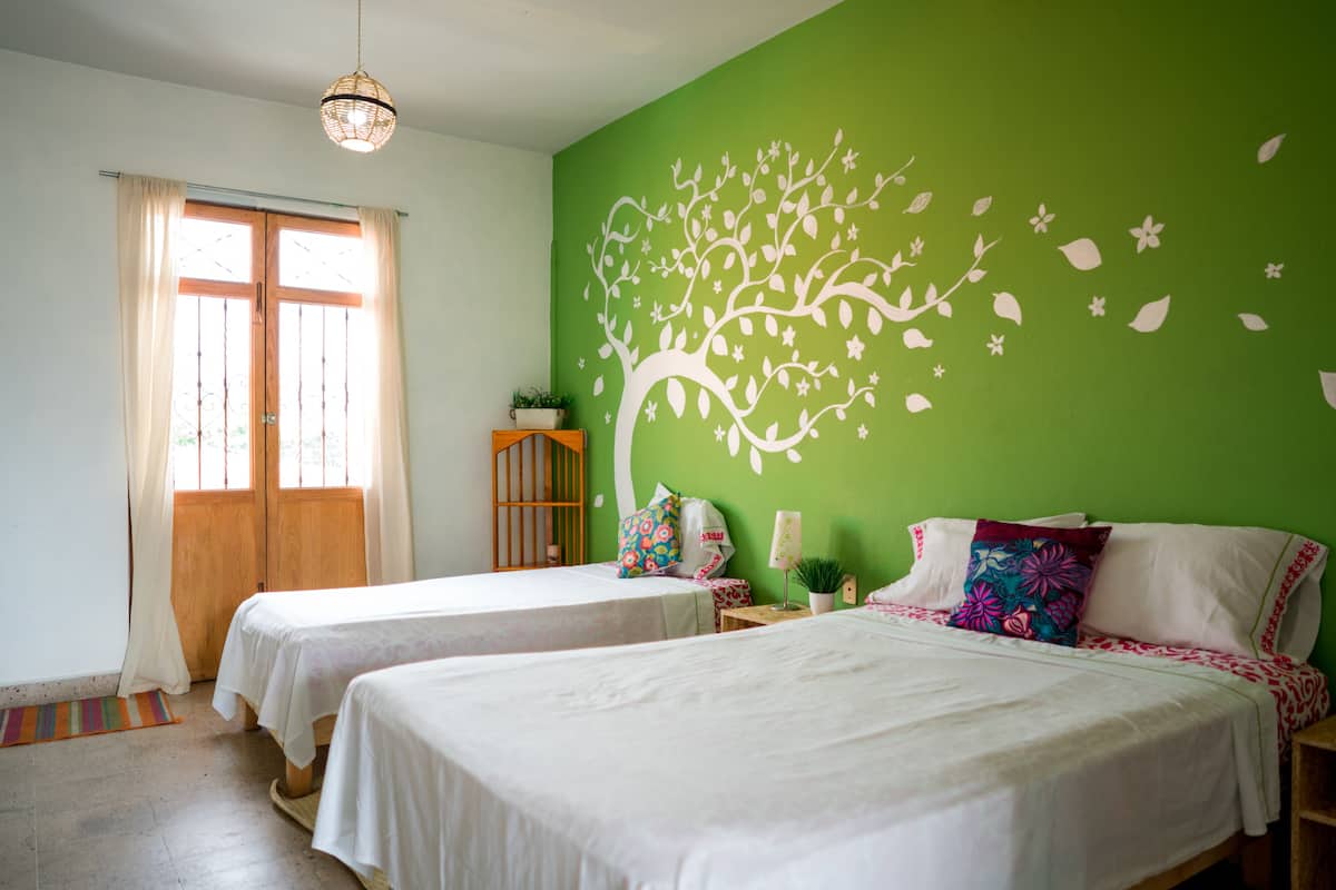 A room with two beds, a green wall with a tree mural, a wooden door, and a small table with a plant.