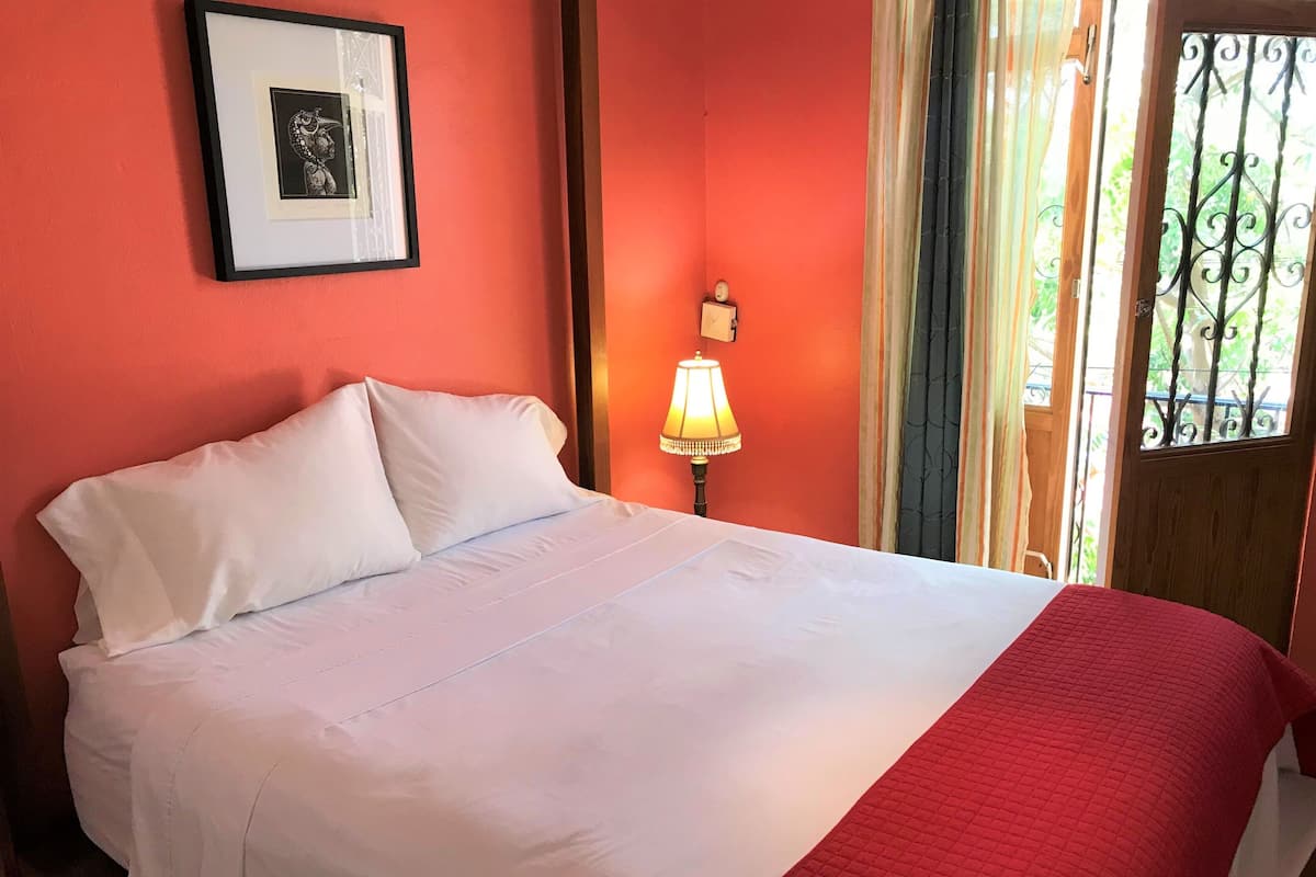 A neatly made bed with white linens and a red blanket, a framed picture on the wall, a bedside lamp, and a window with a view of greenery.