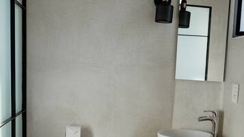 Un baño con inodoro blanco, un lavabo sobre un estante de madera, un espejo sobre el lavabo y una cesta de mimbre en el suelo.