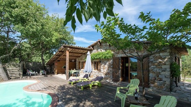 Une terrasse en bois avec une piscine, un espace salon extérieur et un bâtiment en pierre surmonté d’une pergola.
