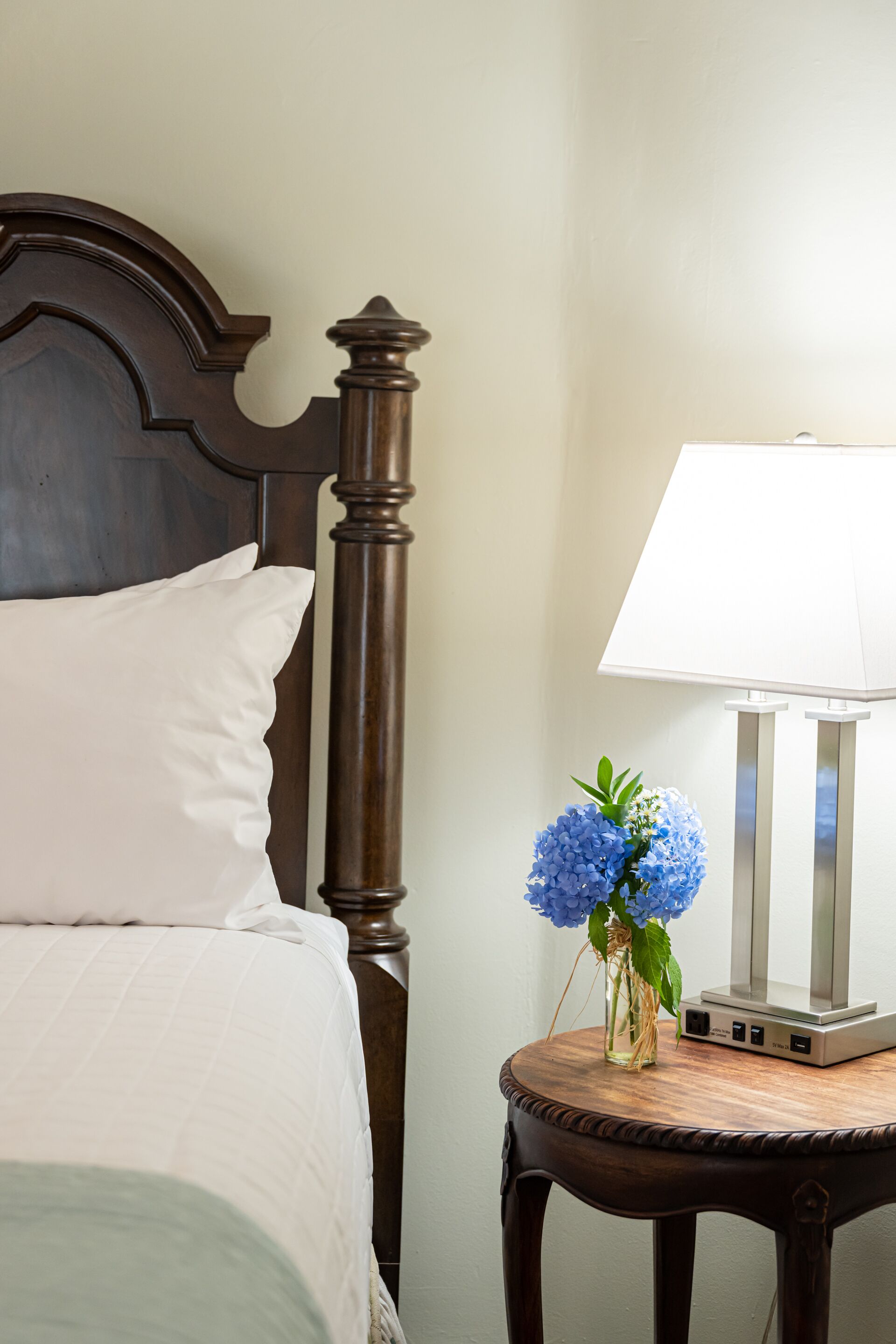 Un lit bien fait, avec un oreiller blanc et une tête de lit en bois. Une petite table en bois, sur laquelle se trouvent une lampe blanche et un vase de fleurs bleues, est placée à côté du lit.