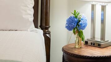 Un lit bien fait, avec un oreiller blanc et une tête de lit en bois. Une petite table en bois, sur laquelle se trouvent une lampe blanche et un vase de fleurs bleues, est placée à côté du lit.