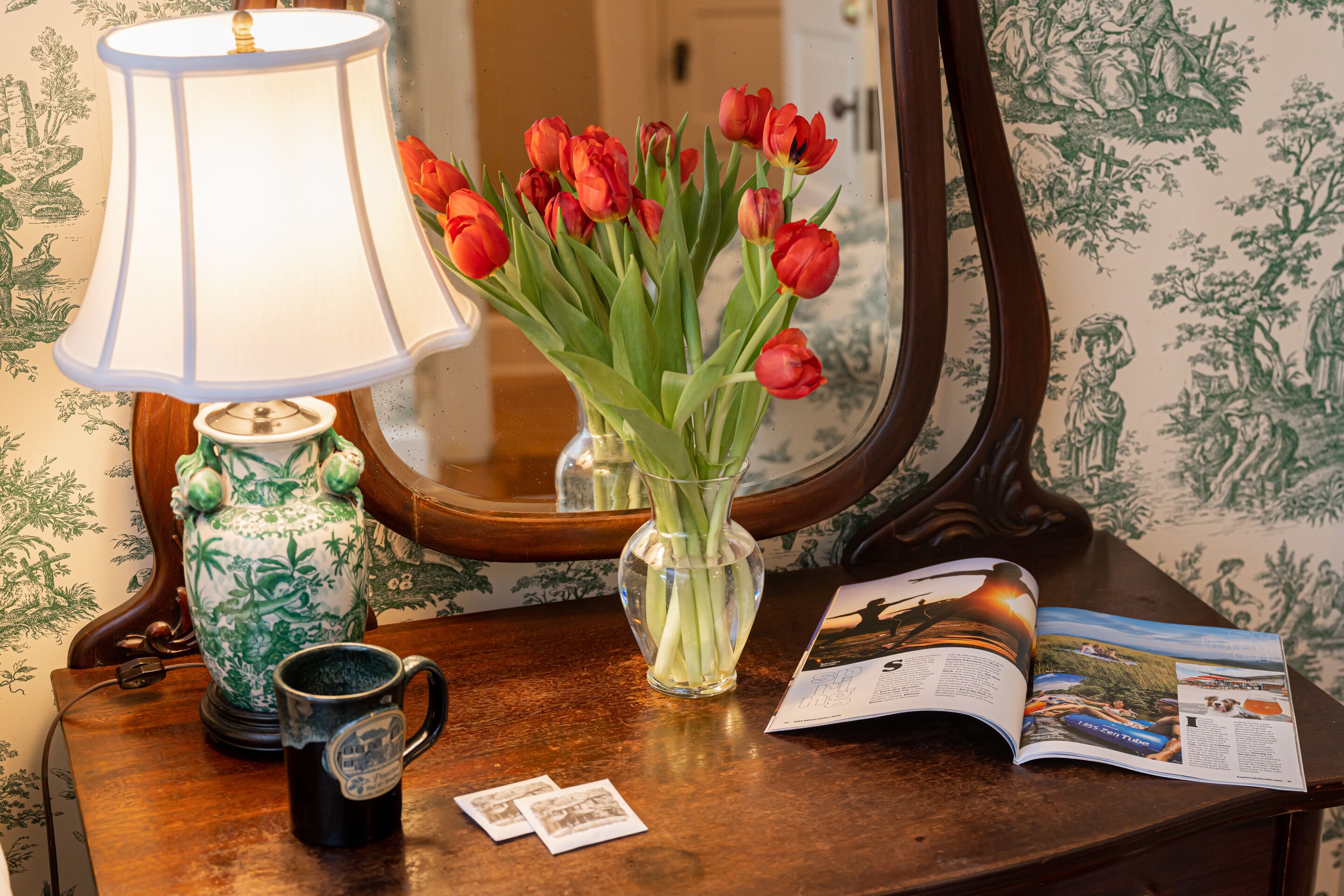 A table with a lamp, a vase of red tulips, a mug, and a magazine.