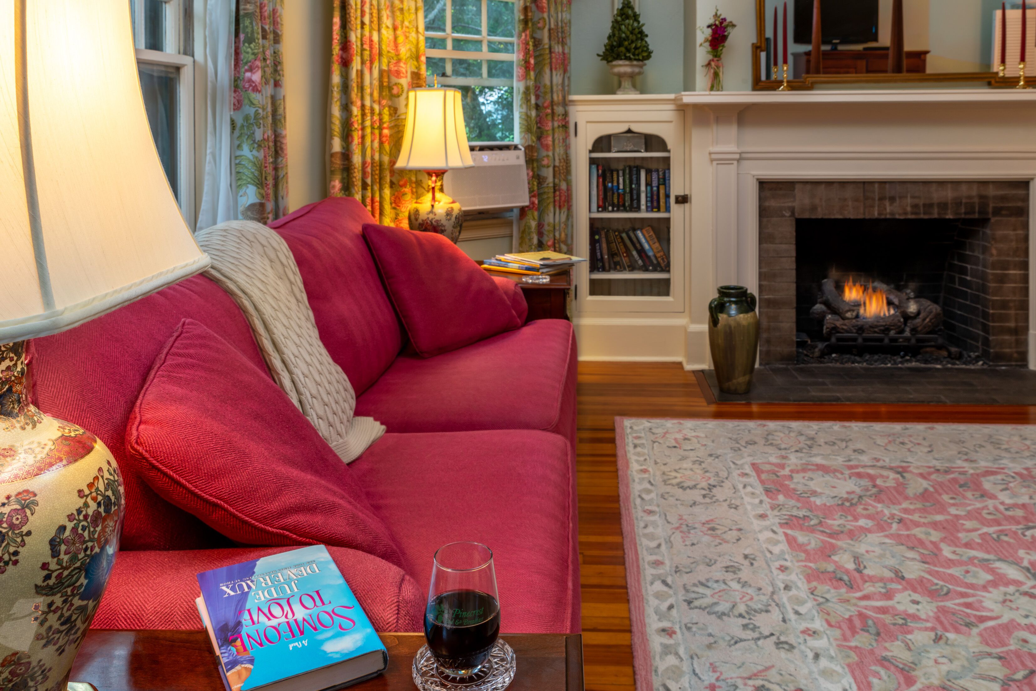 A cozy living room with a red sofa, a fireplace, a bookshelf, and a table with a book and a glass of wine.
