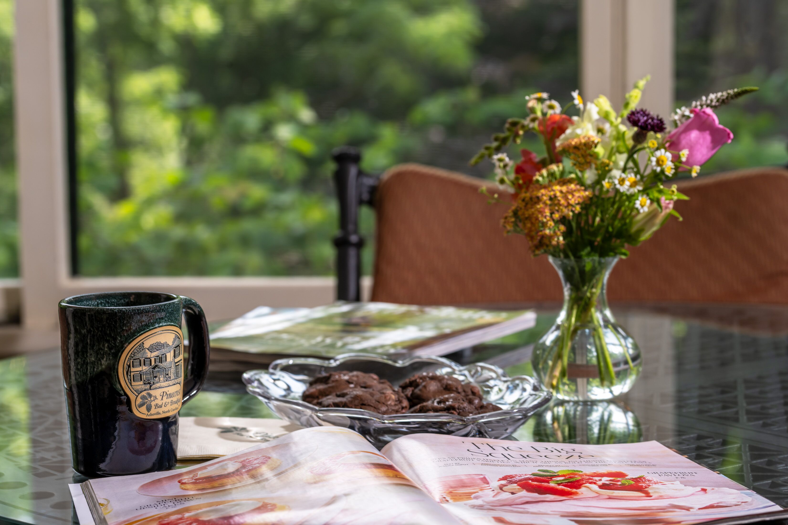 Een tafel met een koffiemok, een glazen schaal vol koekjes, een bloevas en een tijdschrift.