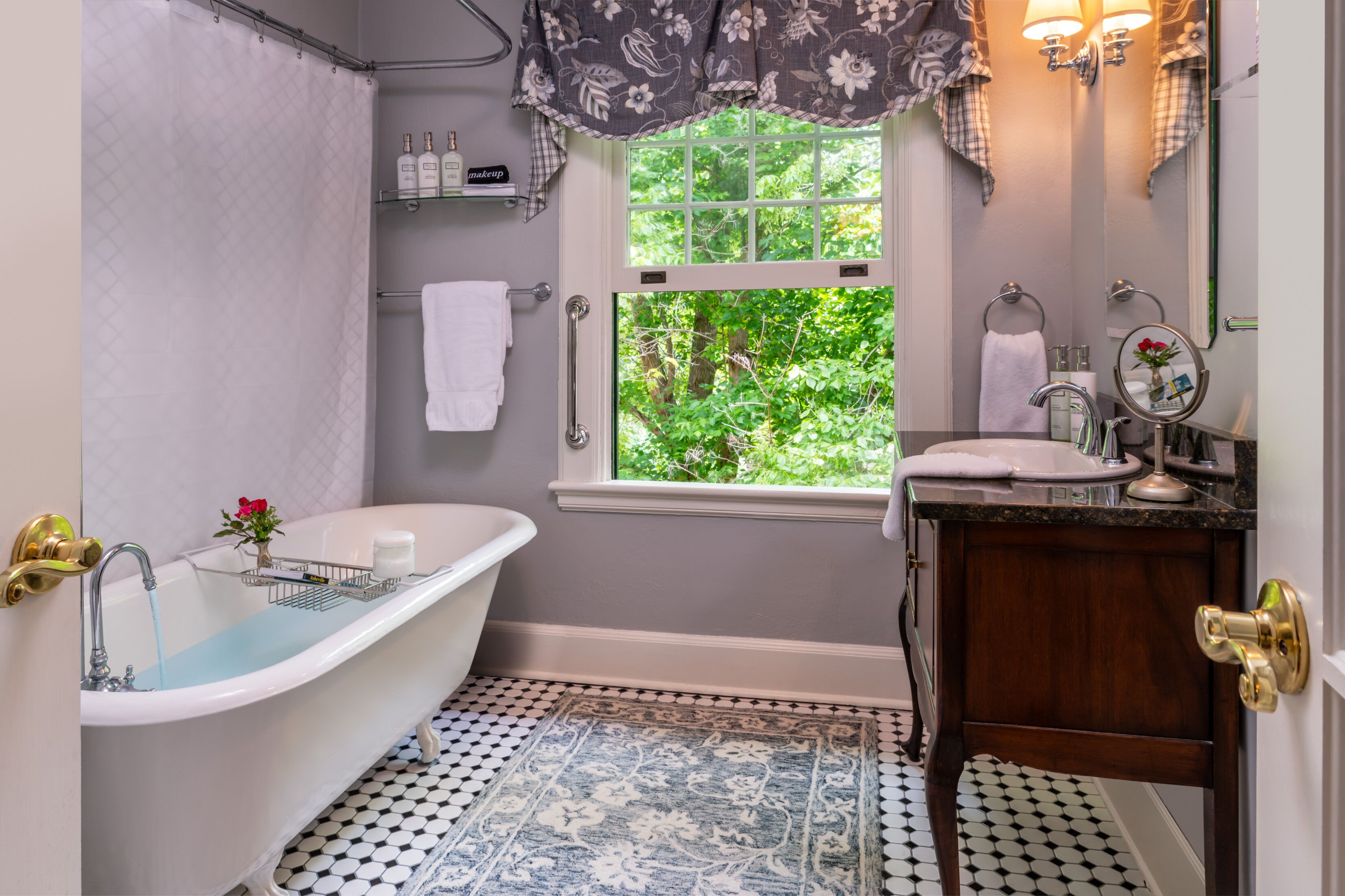 A bathroom with a freestanding bathtub, a window with a view of greenery, a sink with a mirror, and a patterned floor.