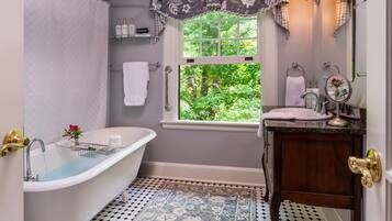 A bathroom with a freestanding bathtub, a window with a view of greenery, a sink with a mirror, and a patterned floor.