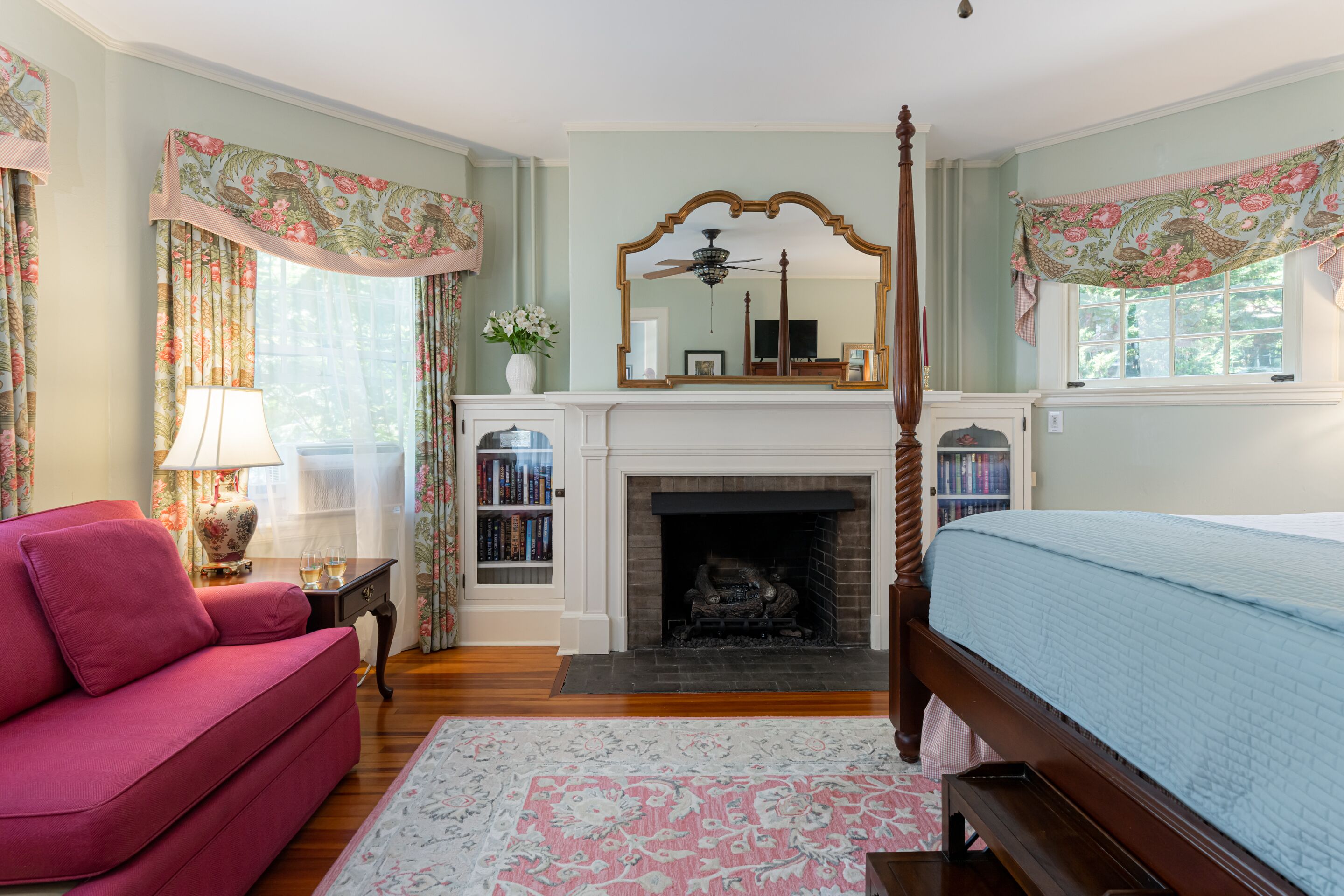 A bedroom with a four-poster bed, a fireplace, a bookshelf, and a red armchair.