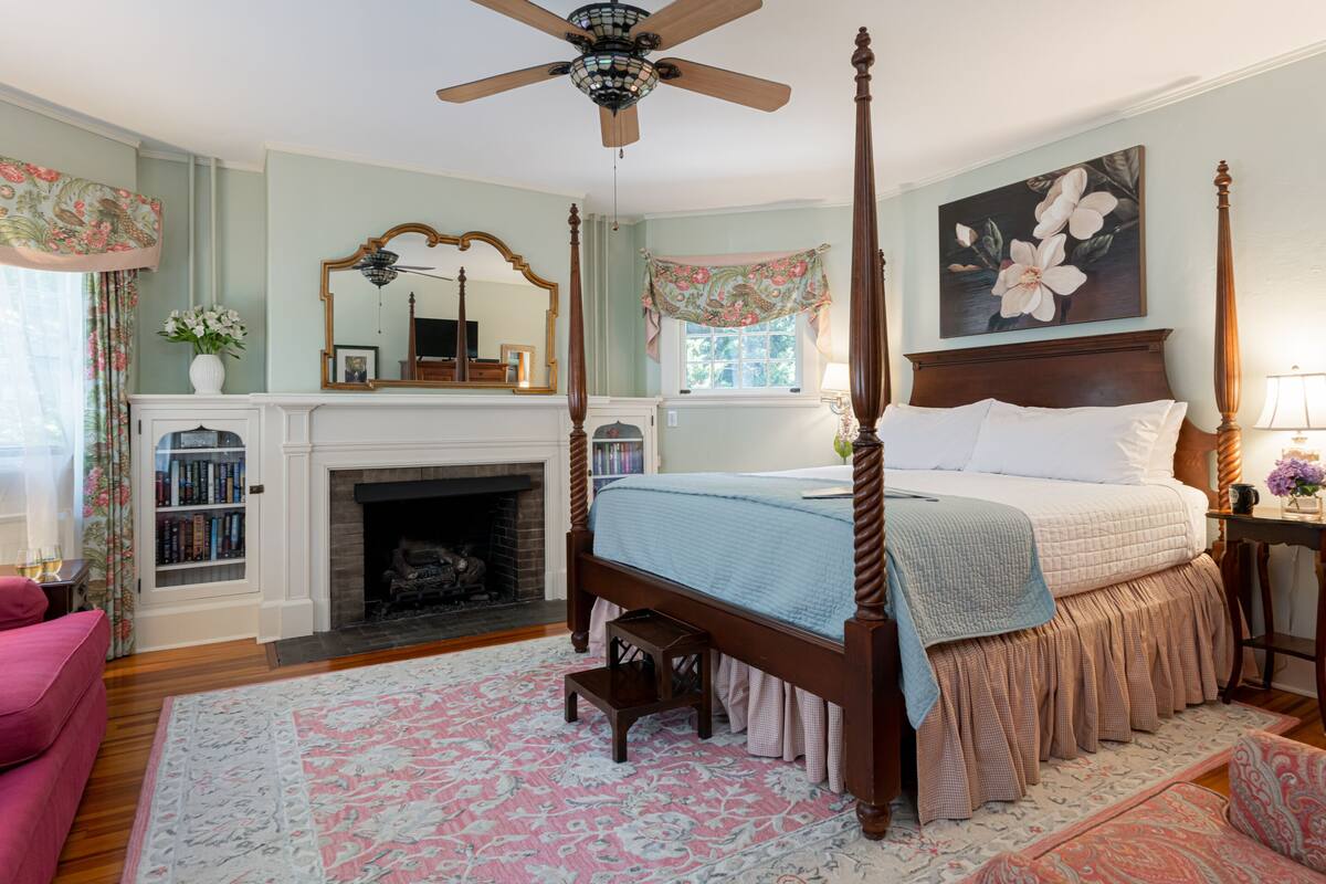 A bedroom with a four-poster bed, a fireplace, a bookshelf, and a large mirror.