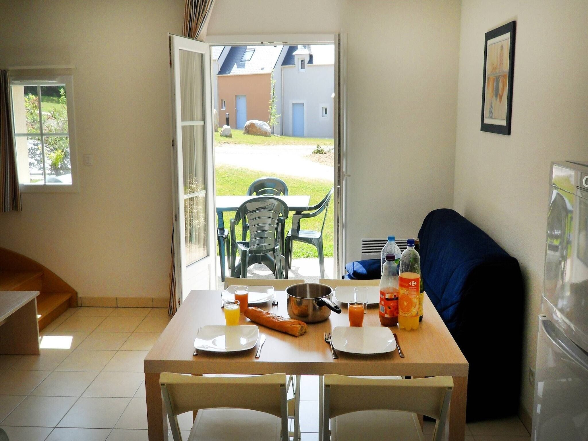 A dining area with a table set for two, a sofa, and a view of a garden through an open door.