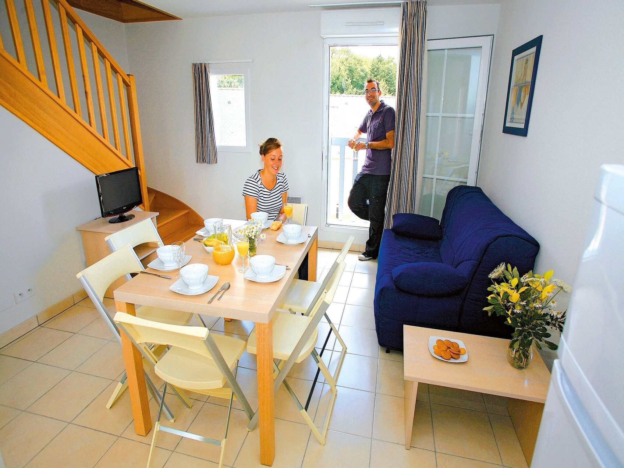 A dining area with a table set for two, a blue sofa, and a small table with a flower vase.