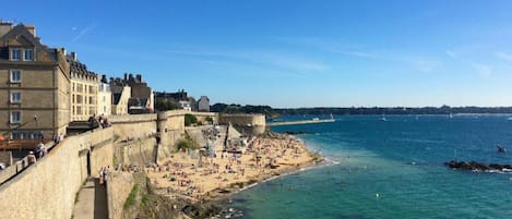 Un pueblo costero con un muro fortificado, una playa de arena y un mar azul cristalino.