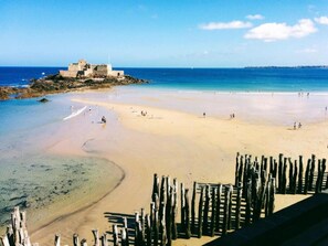 Une plage avec un rivage sablonneux, un petit promontoire rocheux sur lequel se trouve un bâtiment, et une clôture en bois au premier plan.