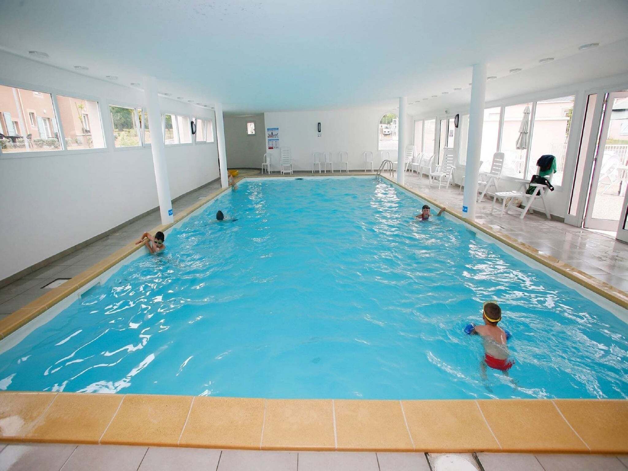 Ein Hallenbad mit klarem Wasser, umgeben von weißen Wänden und Säulen, mit mehreren Sitzbereichen ausgestattet.