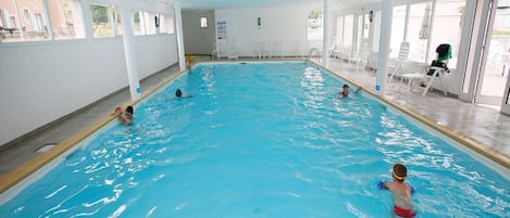 Una piscina interior con agua cristalina, rodeada de paredes y columnas blancas, y equipada con varias zonas de estar.