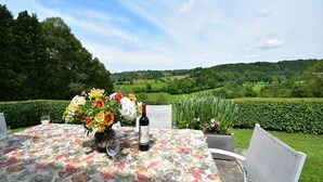 Ein Tisch im Freien mit einer blumigen Tischdecke, einer Weinflasche und Gläsern, vor einer malerischen Kulisse aus sanften Hügeln und blauem Himmel.