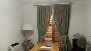 A small room with a wooden table, a white radiator, a fan, a lamp, and a window with green curtains. - Holiday Home in Giardini Naxos near Beach (Taormina Messina)