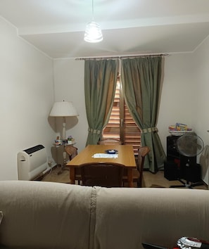 A small room with a wooden table, a white radiator, a fan, a lamp, and a window with green curtains.