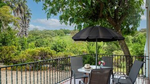 A balcony with a table and chairs, a black umbrella, and a view of a lush garden.