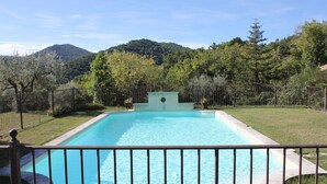 A rectangular swimming pool with a central fountain, surrounded by a metal fence, set against a backdrop of lush greenery and a mountain. - Holiday Home near Mont Ventoux Pool (Vaison-La-Romaine)